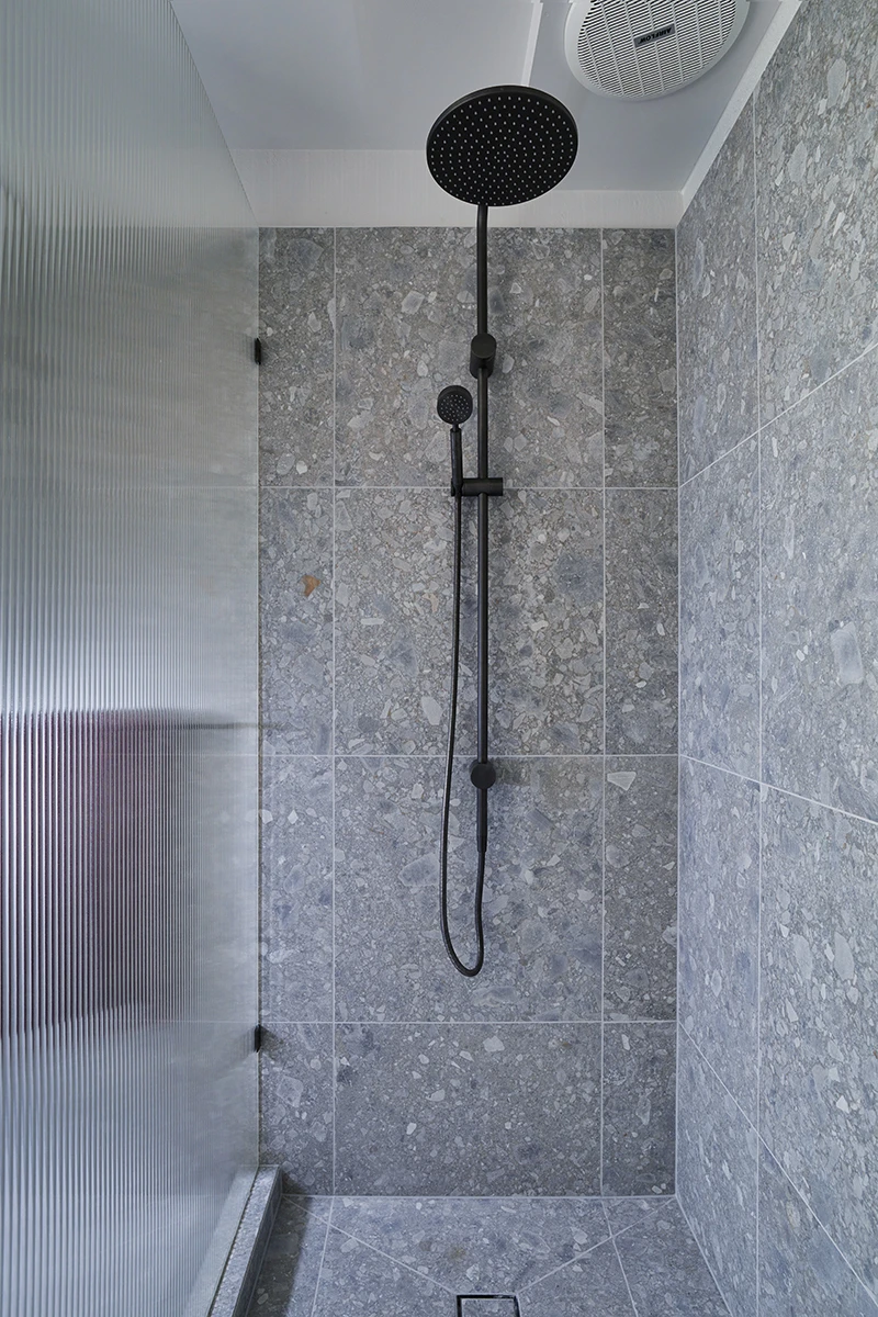 Modern shower with black rainfall showerhead and handheld attachment against gray stone tile walls and glass panel.