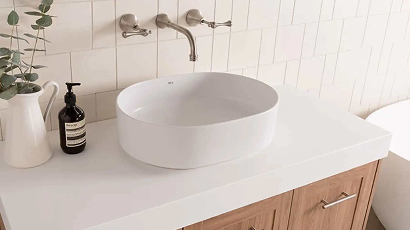 Modern bathroom with white vessel sink, wall-mounted brass faucet, wooden vanity, and decorative plant in white pitcher.