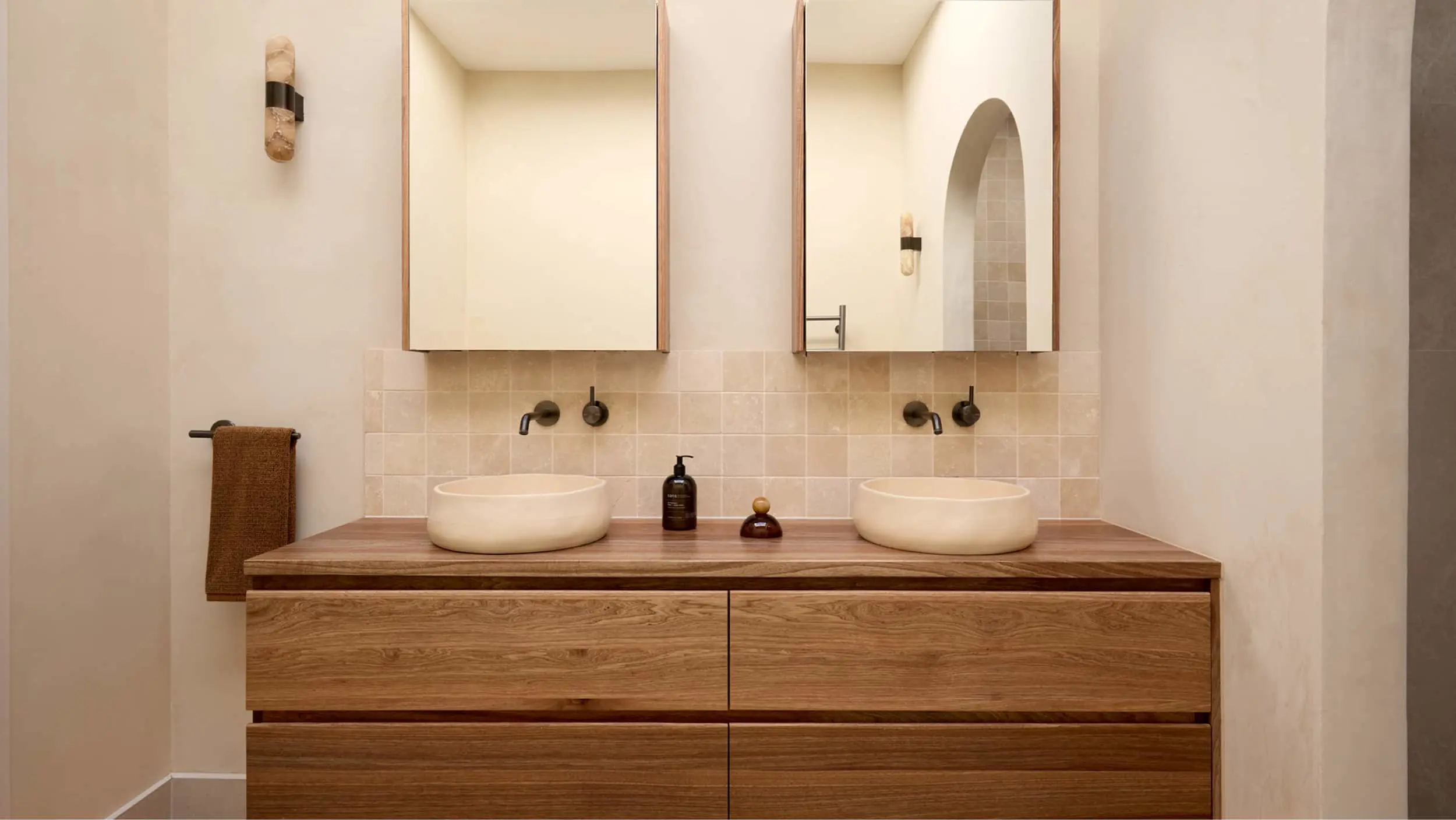 Bathroom vanity with two white vessel sinks on wood counter, two mirrors, beige tile backsplash, and black faucets.