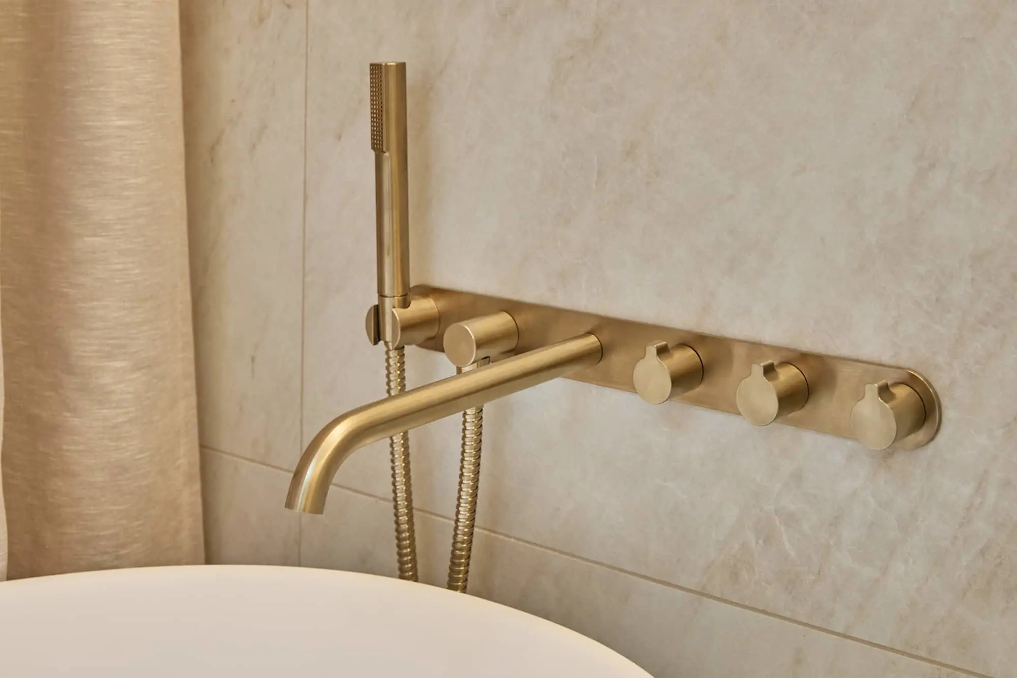 Brushed gold wall-mounted bath filler with three knob controls, a spout, and a handheld shower head on a marble-tiled wall.