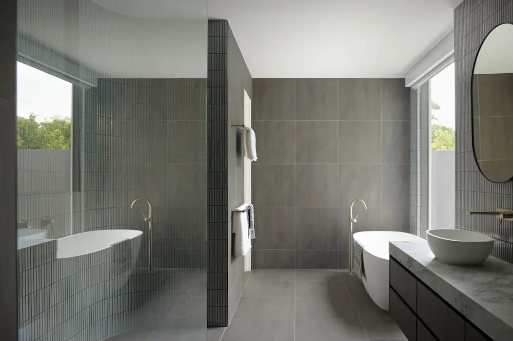 Modern bathroom with gray tile walls, freestanding tub, vessel sink, and round mirror with views of greenery outside.