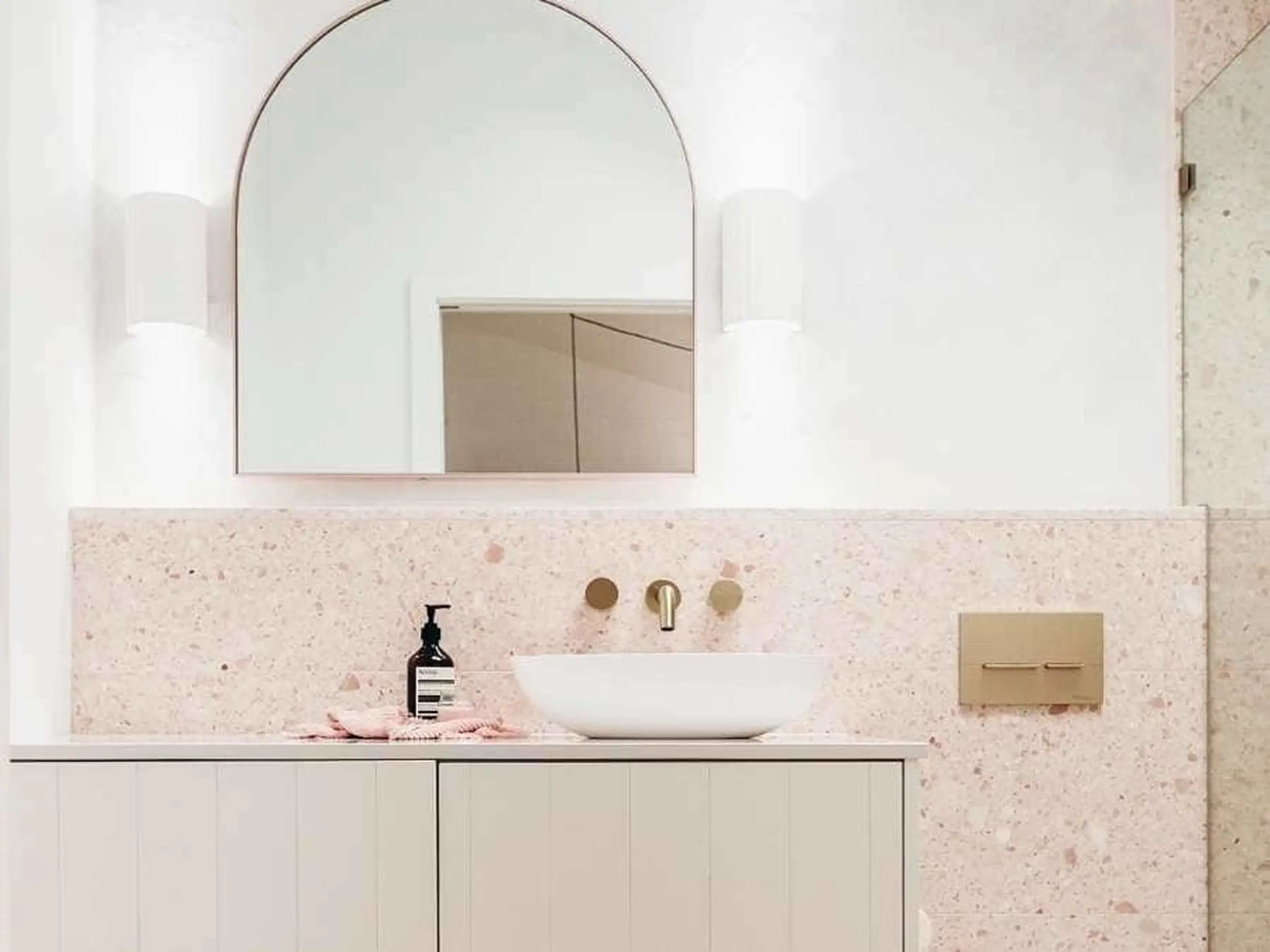 Modern bathroom with arched mirror, white vessel sink, pink terrazzo countertop, and brass fixtures.
