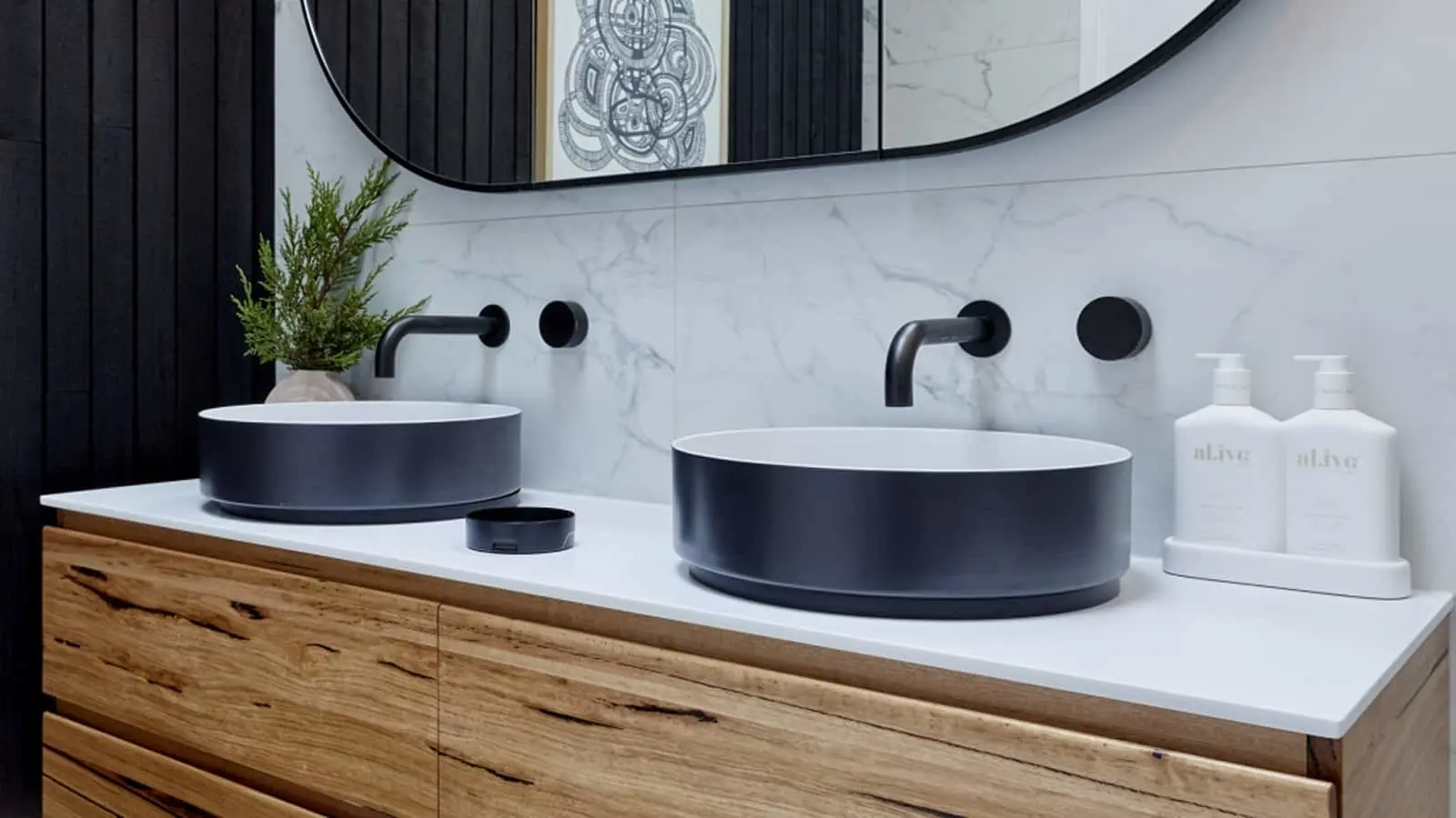 Modern bathroom with two black vessel sinks on wooden vanity, marble backsplash, black faucets, and oval mirror.