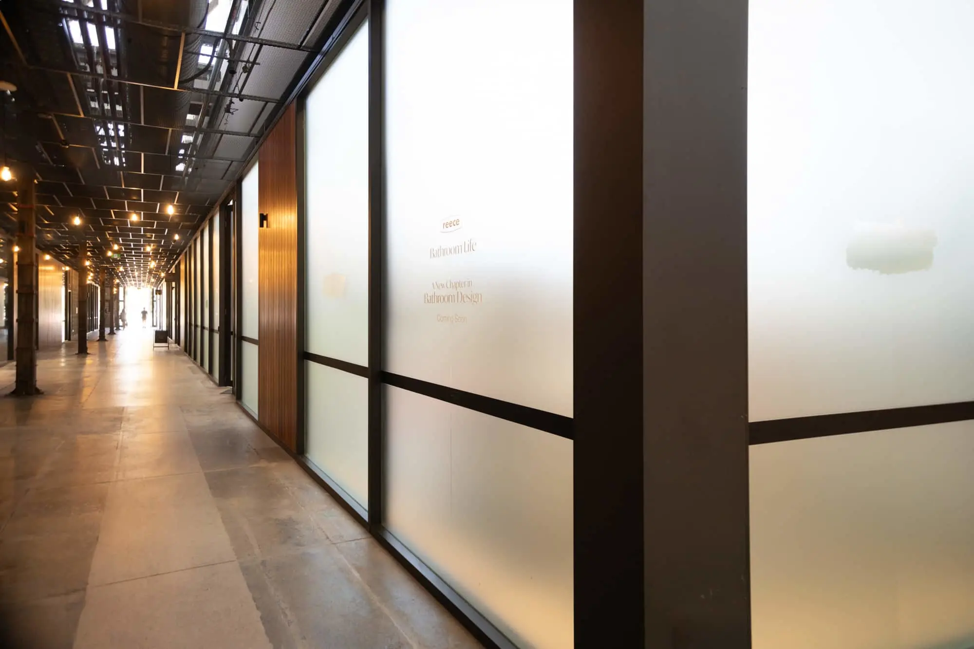 Modern hallway with frosted glass walls, wooden accents, and overhead lighting leading to a bright exit.