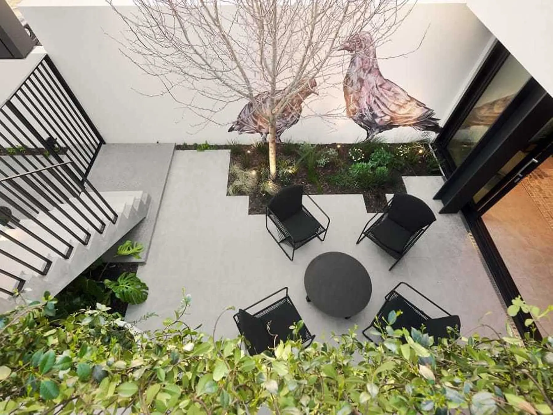 Modern courtyard patio with black chairs around circular table, bare tree, bird artwork, and concrete flooring.