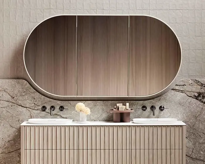 Modern bathroom with oval mirror above double sink vanity, marble wall, and beige cabinetry with vertical grooves.