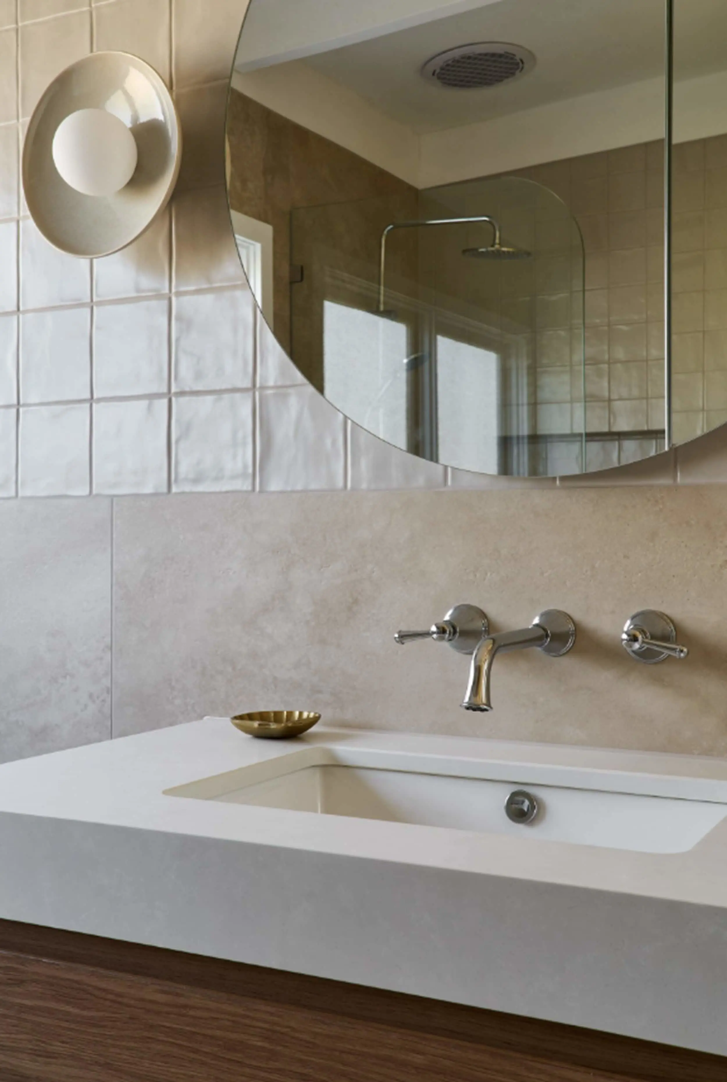 Modern bathroom with white sink, wall-mounted faucet, round mirror, and circular wall light against beige tiled walls.