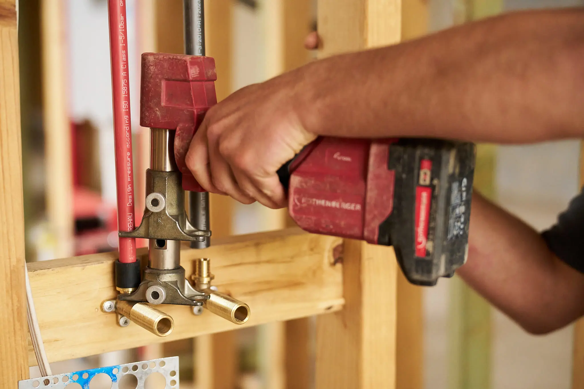 Hand using red power drill to install plumbing pipes and fittings on wooden frame with red PEX tubing.