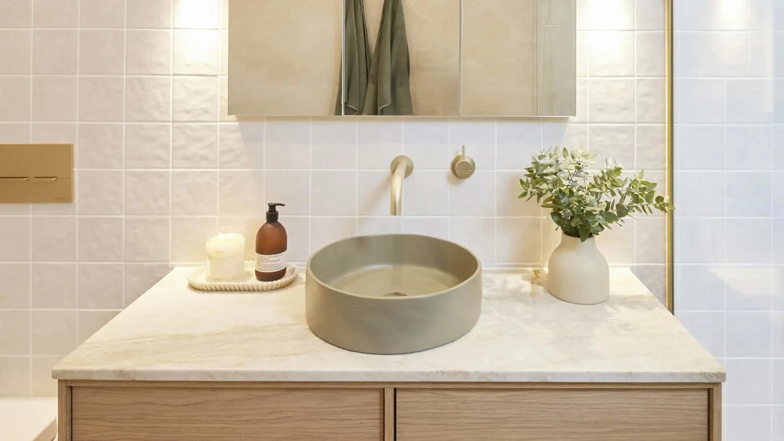 Modern bathroom with round sage basin on marble countertop, wall-mounted brass faucet, soap dispenser, candle, and plant.
