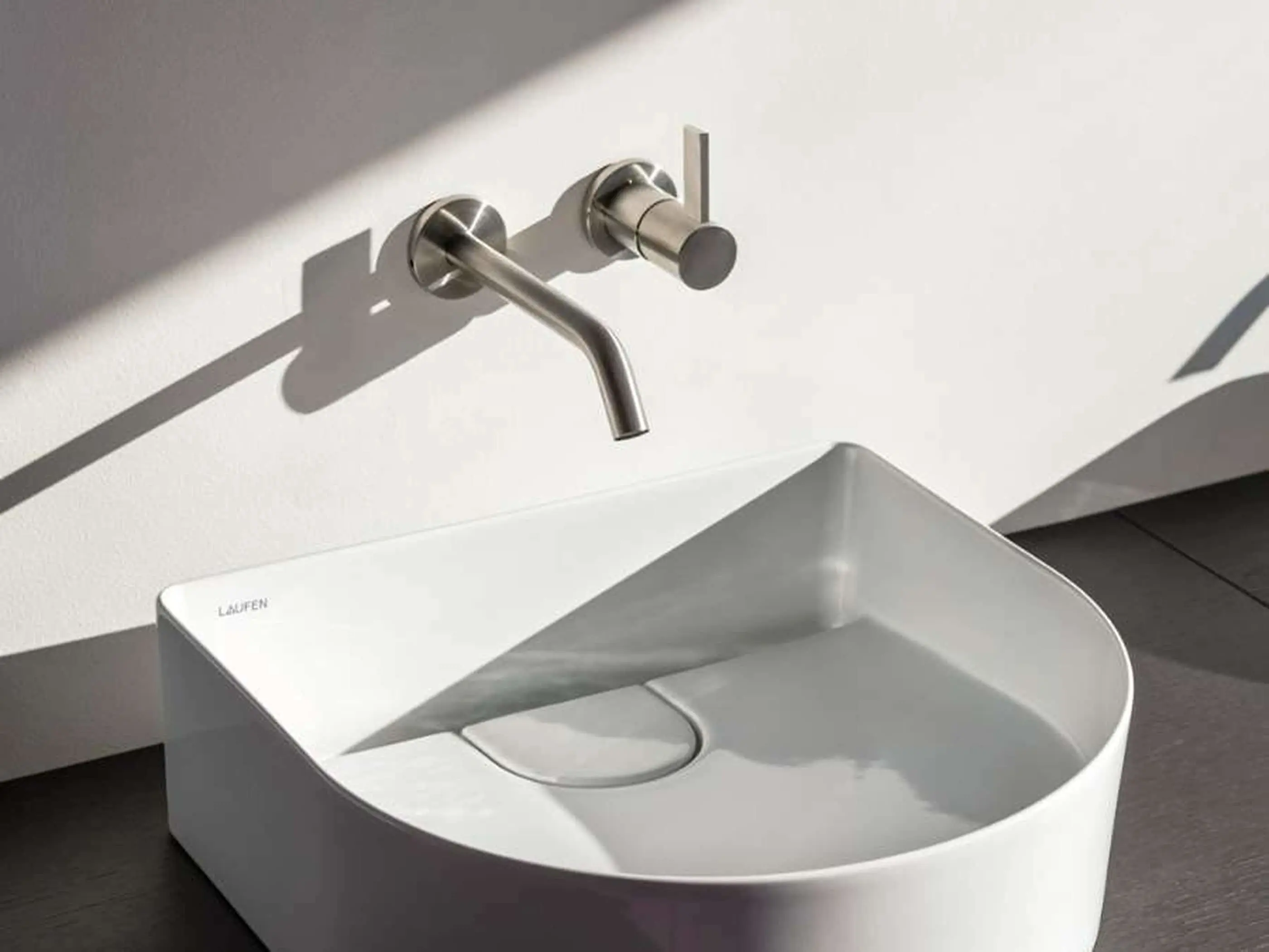 Modern white bathroom sink with wall-mounted brushed steel faucet in minimalist design by Laufen.