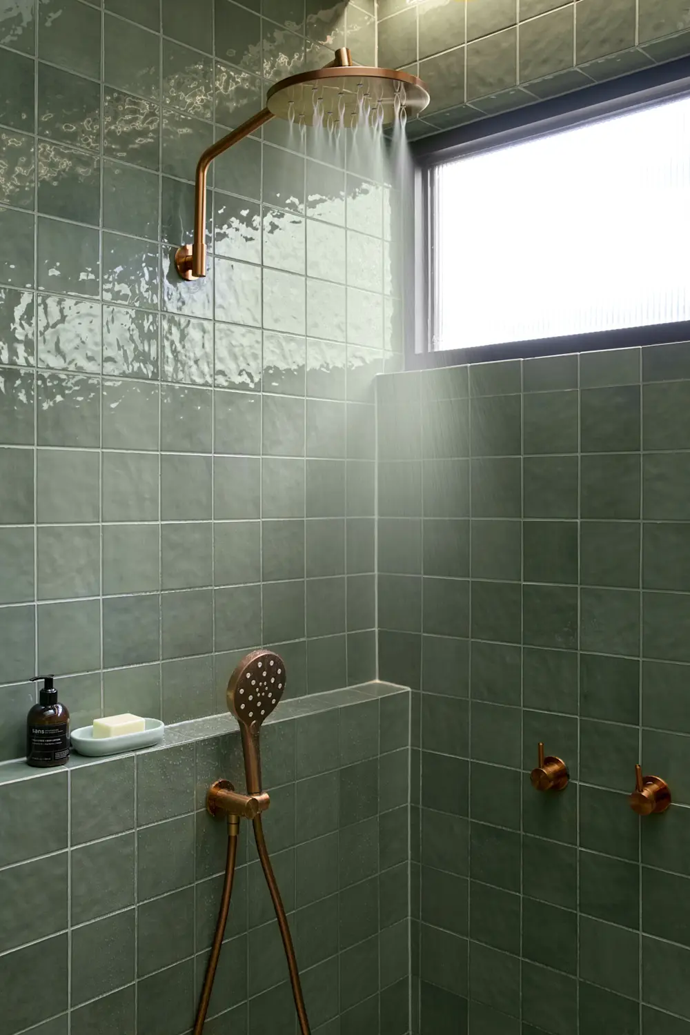 Modern shower with green tile walls, copper fixtures including rainfall showerhead and handheld sprayer, and built-in shelf.