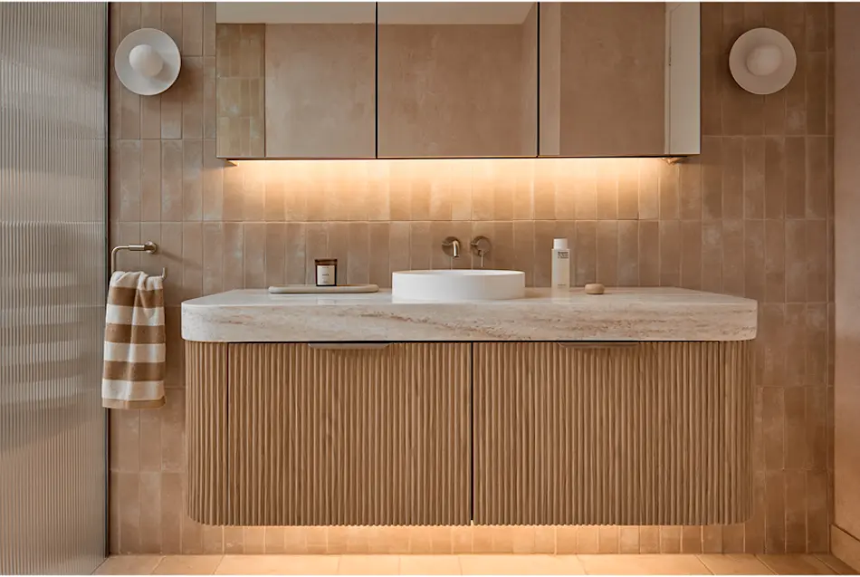 Modern bathroom with beige tiled walls, wooden vanity with stone countertop, white vessel sink, and under-cabinet lighting.
