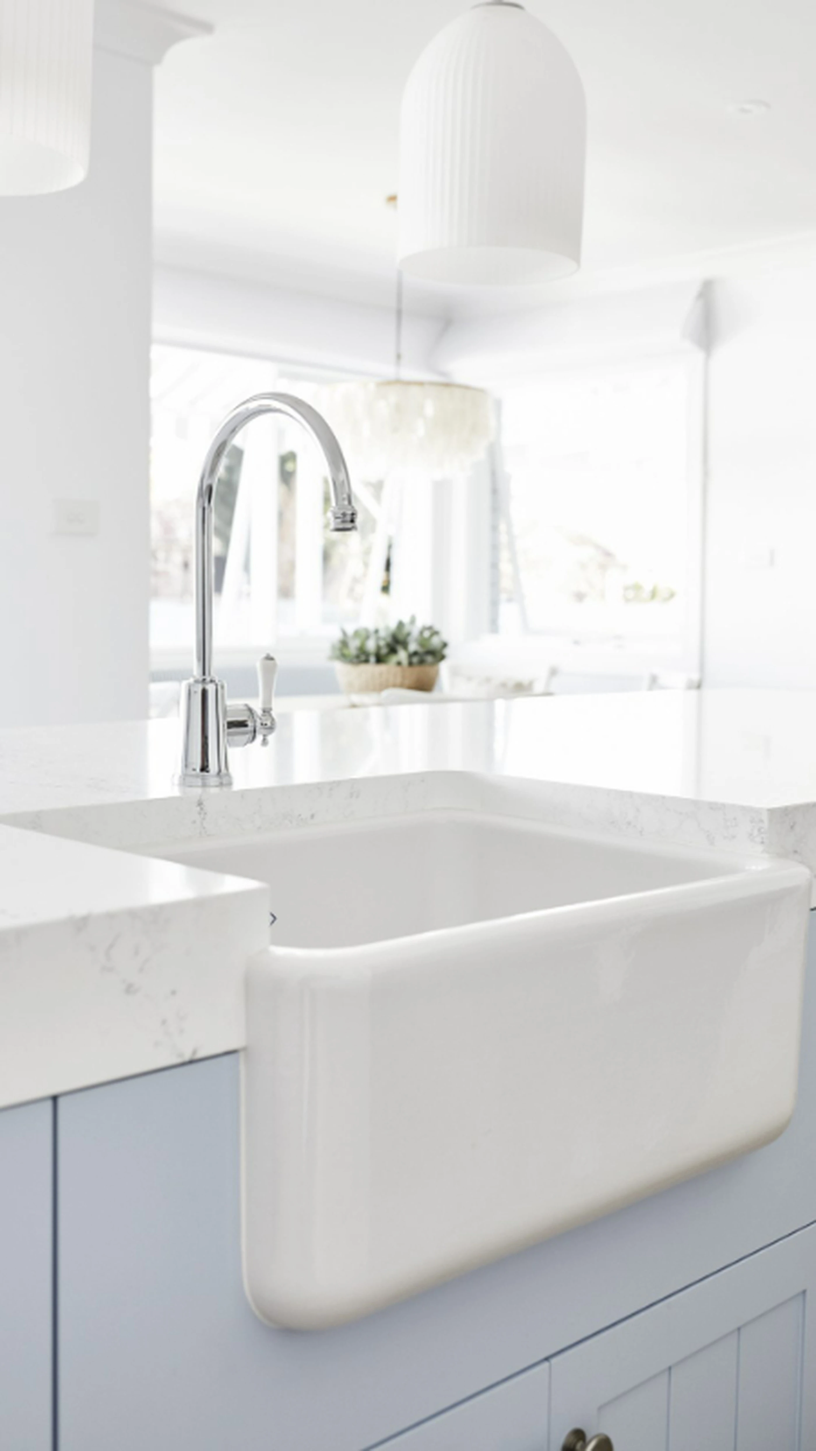 White farmhouse sink with chrome faucet in bright kitchen featuring marble countertops and pendant lighting.