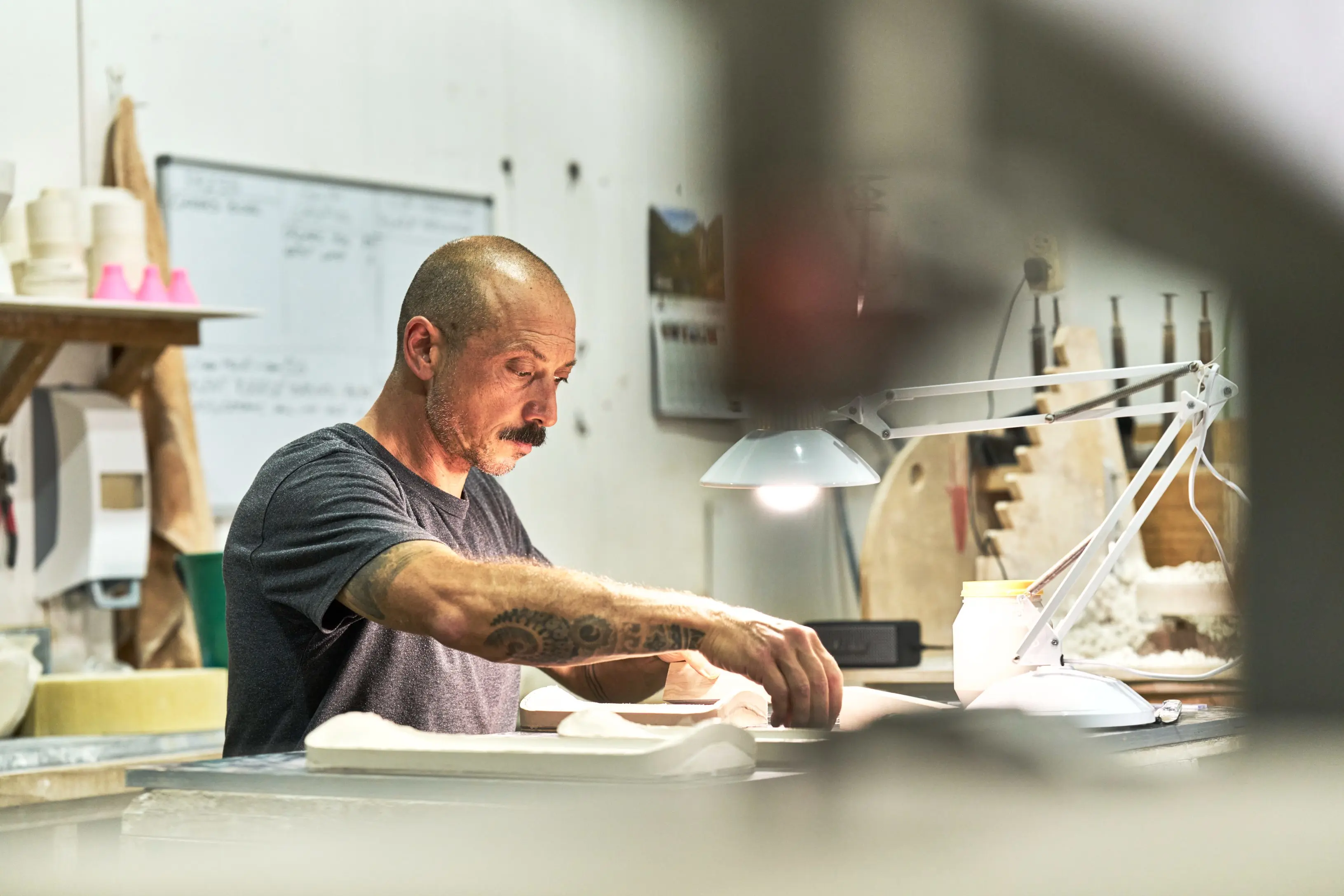 A tattooed man in a grey t-shirt works at a cluttered workshop bench under a white desk lamp.