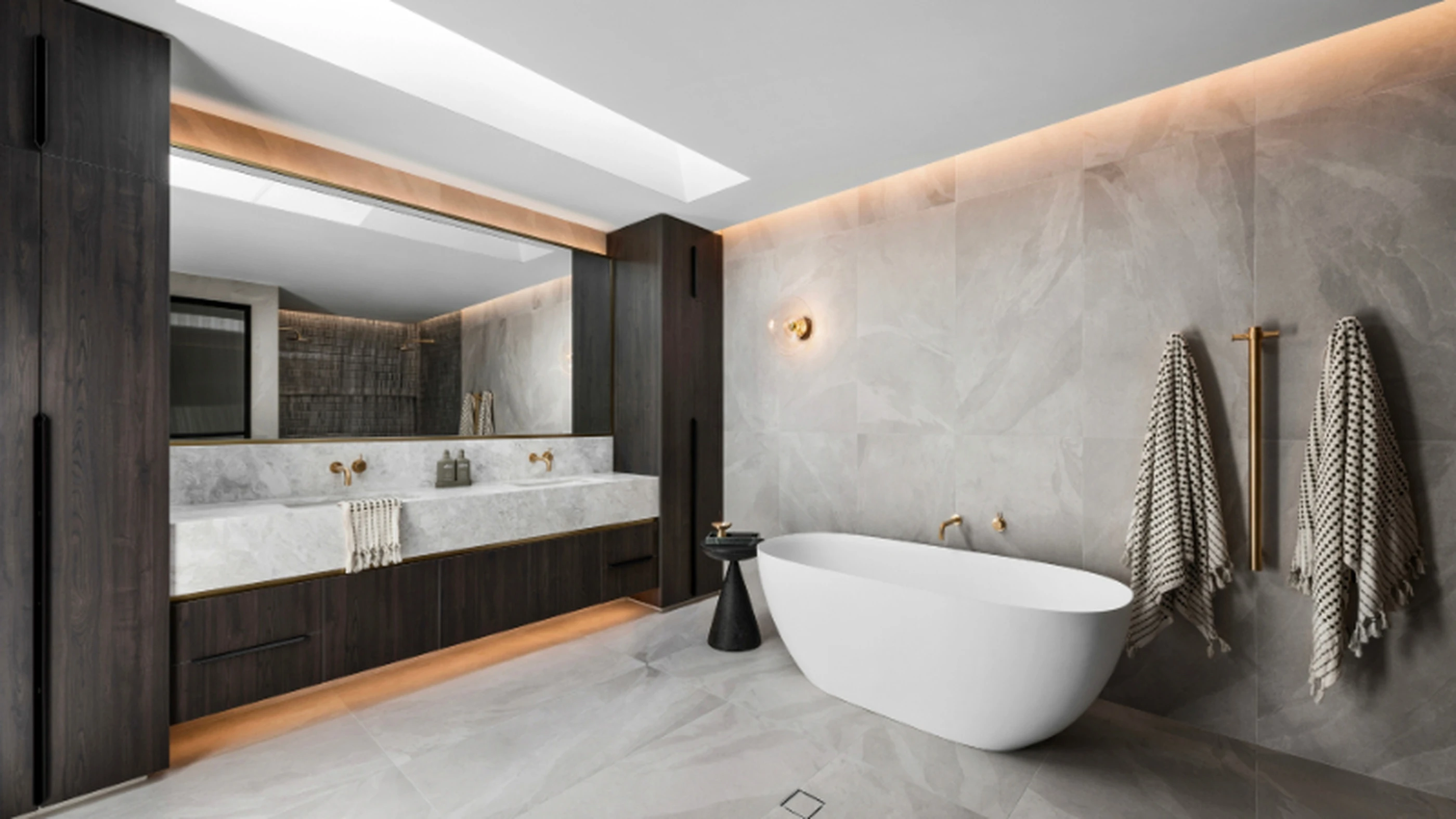 Modern luxury bathroom with white freestanding tub, marble walls, dark wood vanity, and gold fixtures.