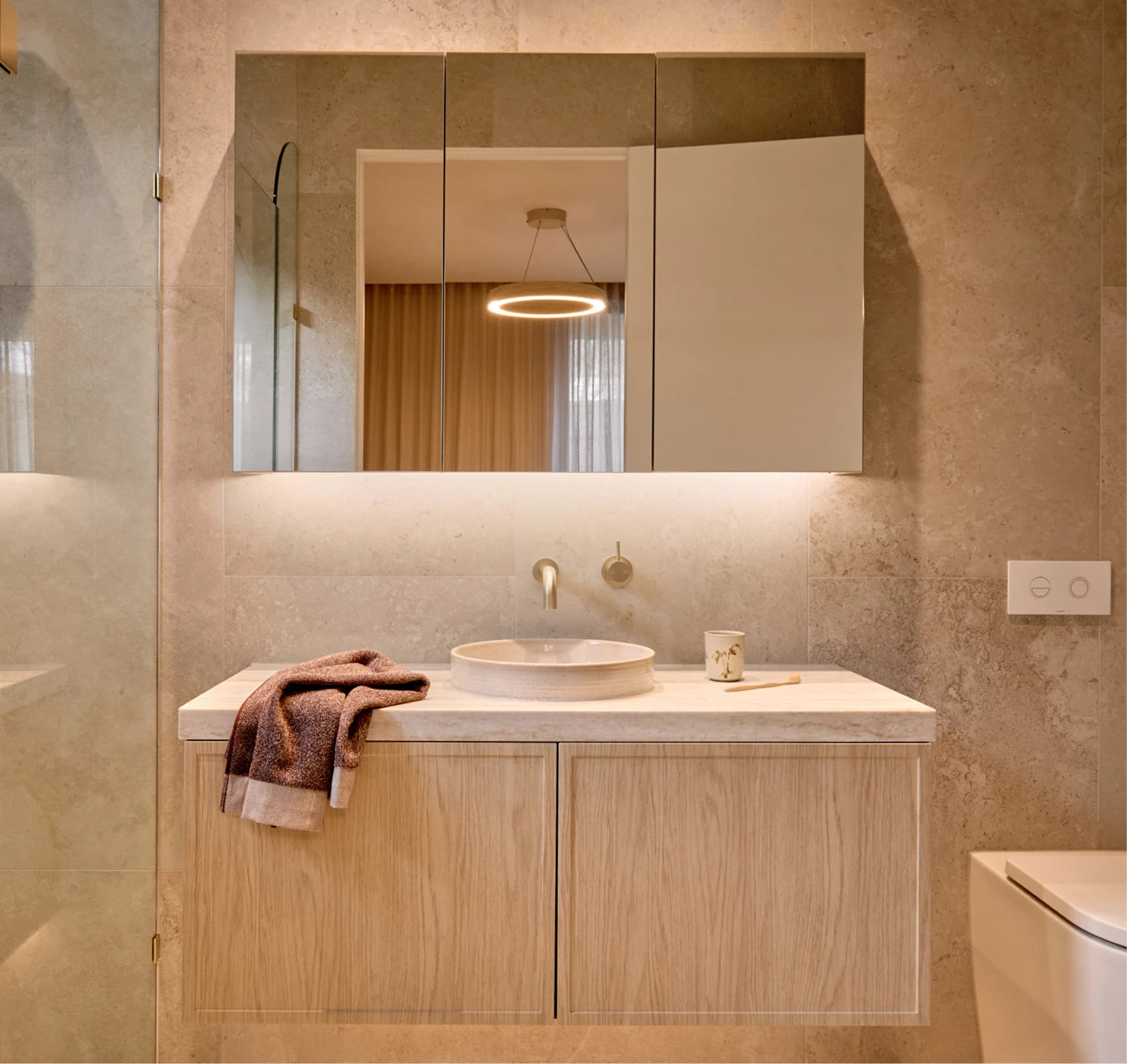 Modern bathroom with beige stone walls, floating wood vanity, round white sink, and backlit mirror cabinet above.