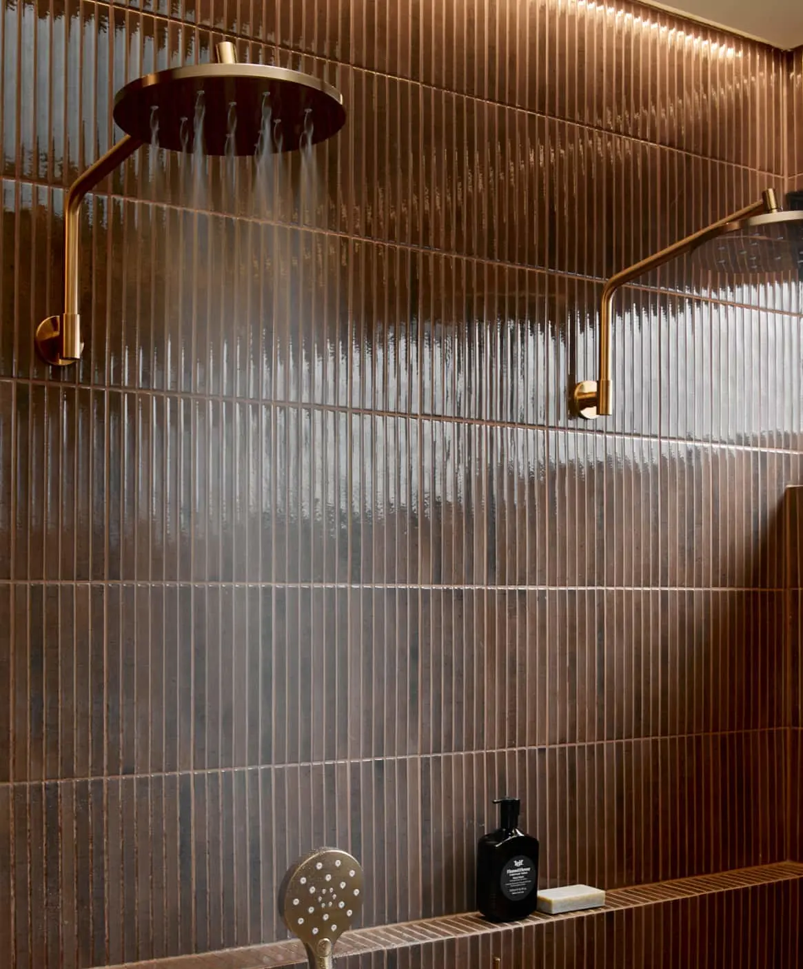 Modern bathroom with bronze tiled walls, rainfall showerhead, and handheld shower fixture with water droplets.
