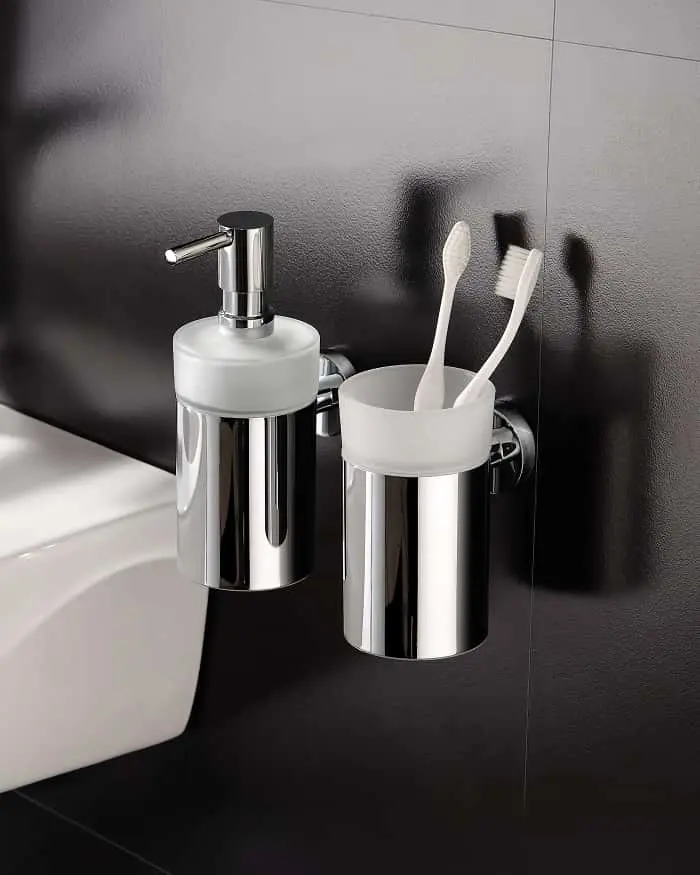 Wall-mounted chrome soap dispenser and toothbrush holder with white tops against a dark textured wall