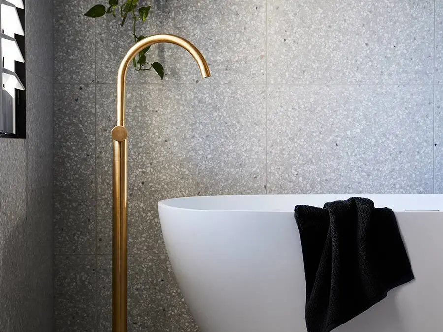 Modern bathroom with white freestanding tub, brass floor-mounted faucet, black towel, and gray stone walls.