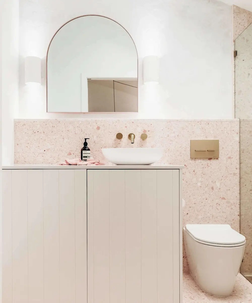 Modern bathroom with pink terrazzo walls, white vanity, vessel sink, arched mirror, wall sconces, and white toilet.