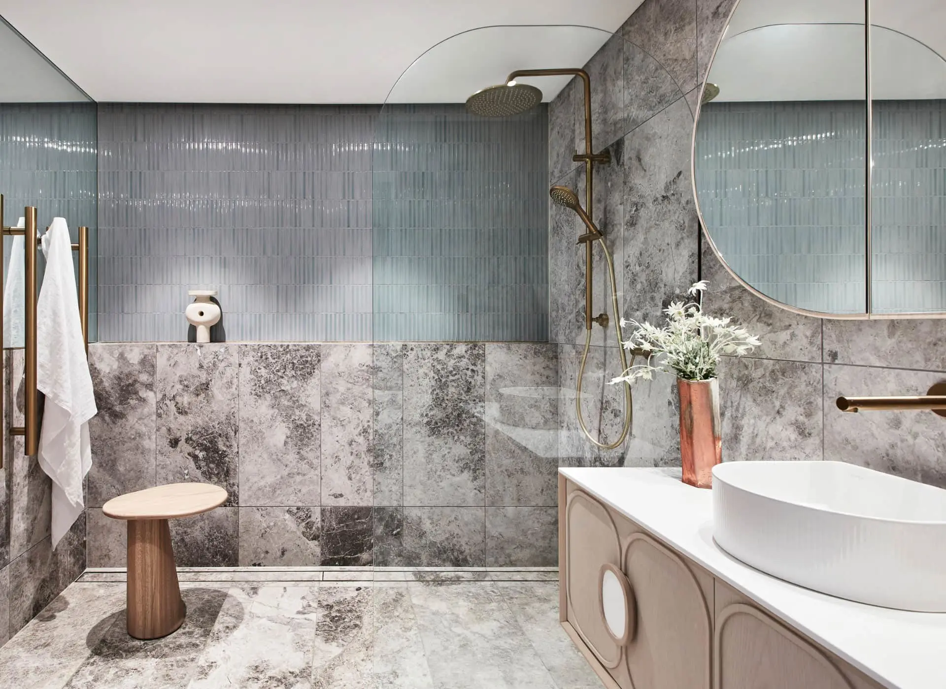 Modern bathroom with blue and marble tiles, brass shower fixtures, round mirror, white sink, and wooden stool.