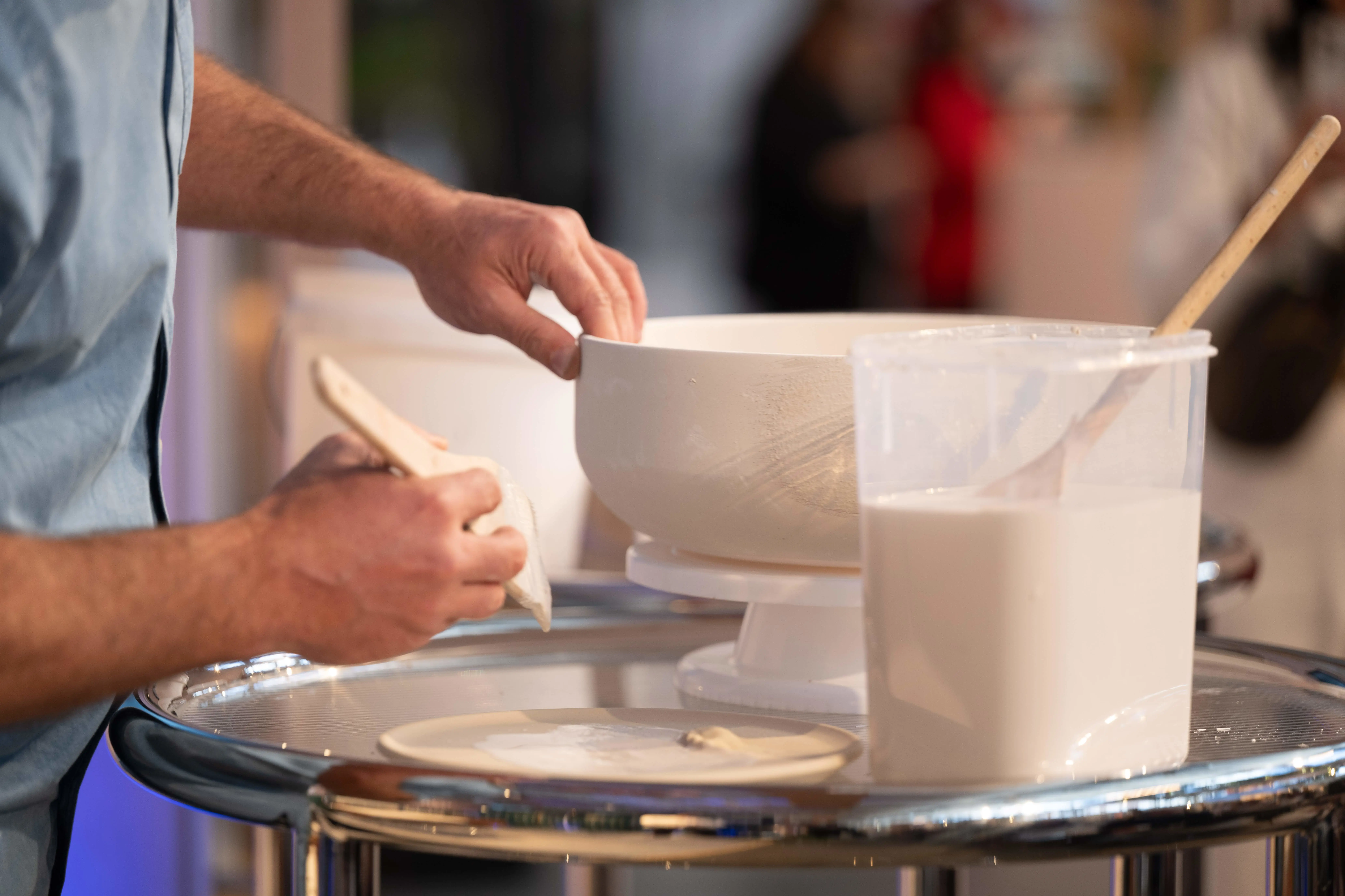 Person shaping pottery on a wheel with hands, white clay bowl and tools visible on work surface.