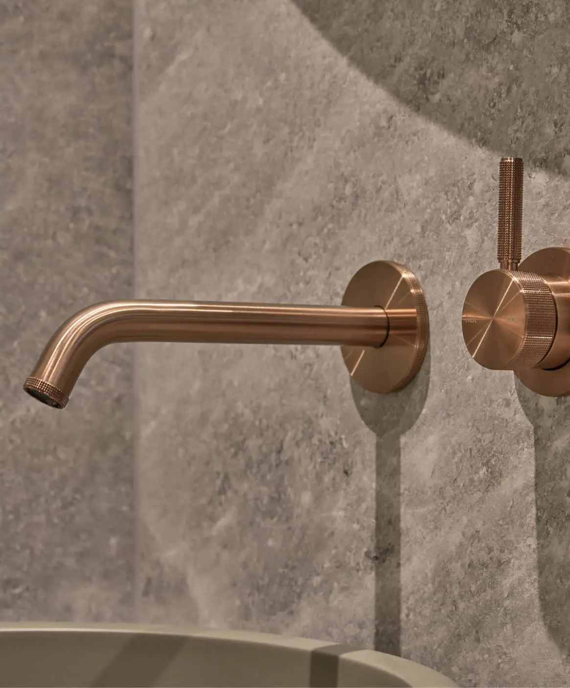 Bronze wall-mounted bathroom faucet with textured handle against gray concrete wall.
