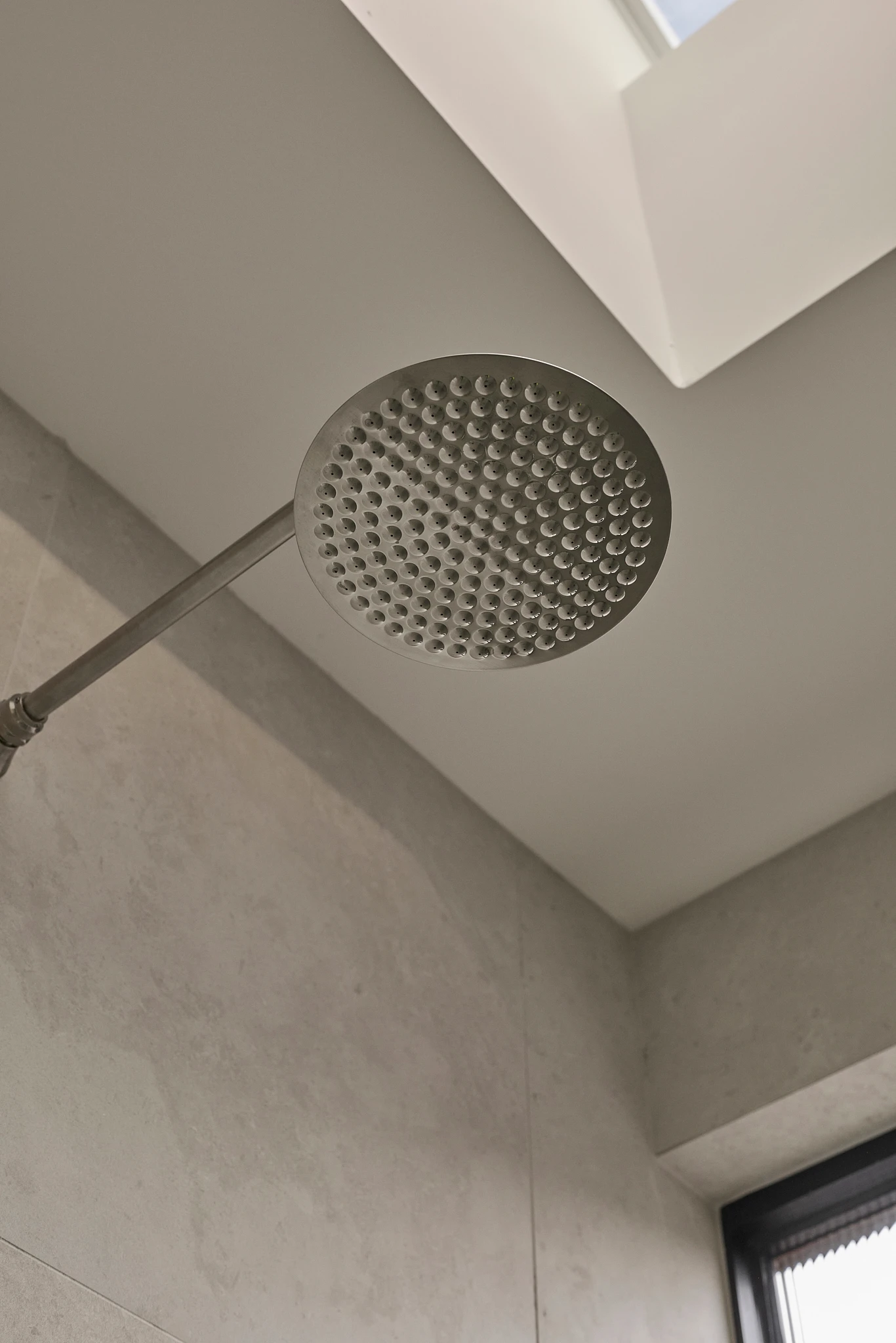 Round stainless steel rainfall showerhead mounted to ceiling in a minimalist bathroom with light beige walls.
