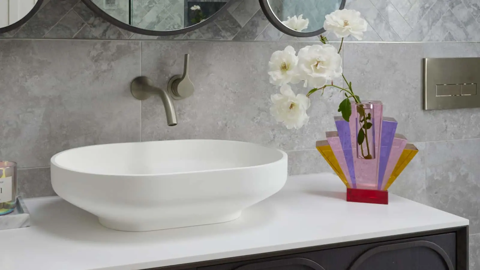 Modern bathroom with white vessel sink, wall-mounted faucet, and colorful art deco vase holding white roses.