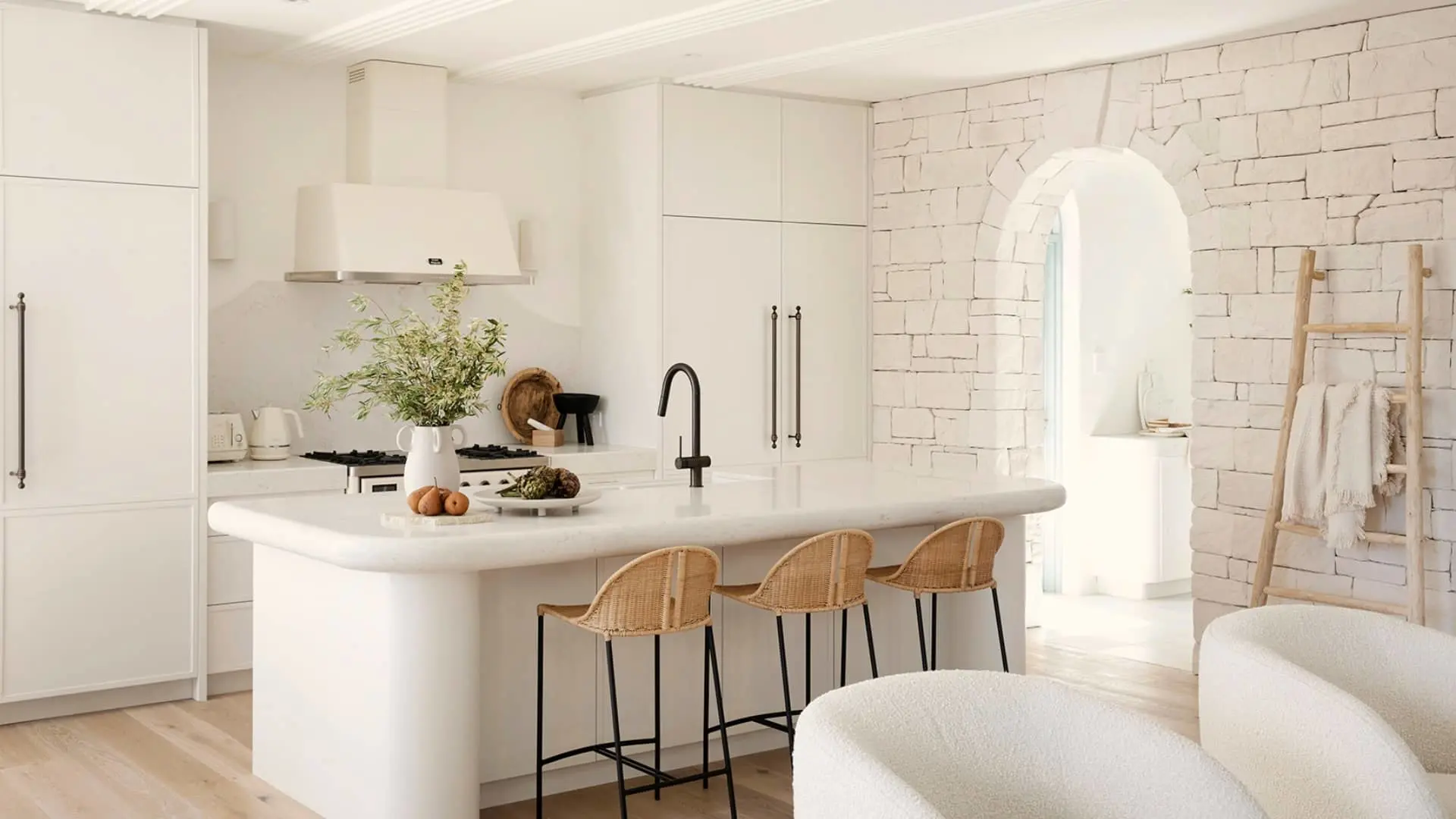 Modern white kitchen with stone wall, arched doorway, rattan barstools, and marble island with plants and fruit.