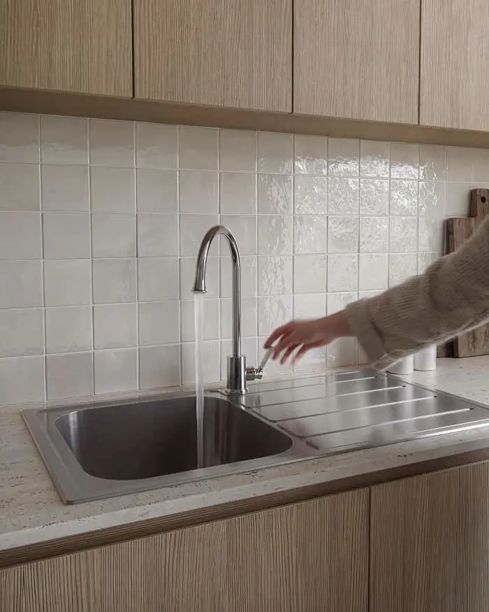 Hand reaching toward stainless steel kitchen sink with running water and white tile backsplash