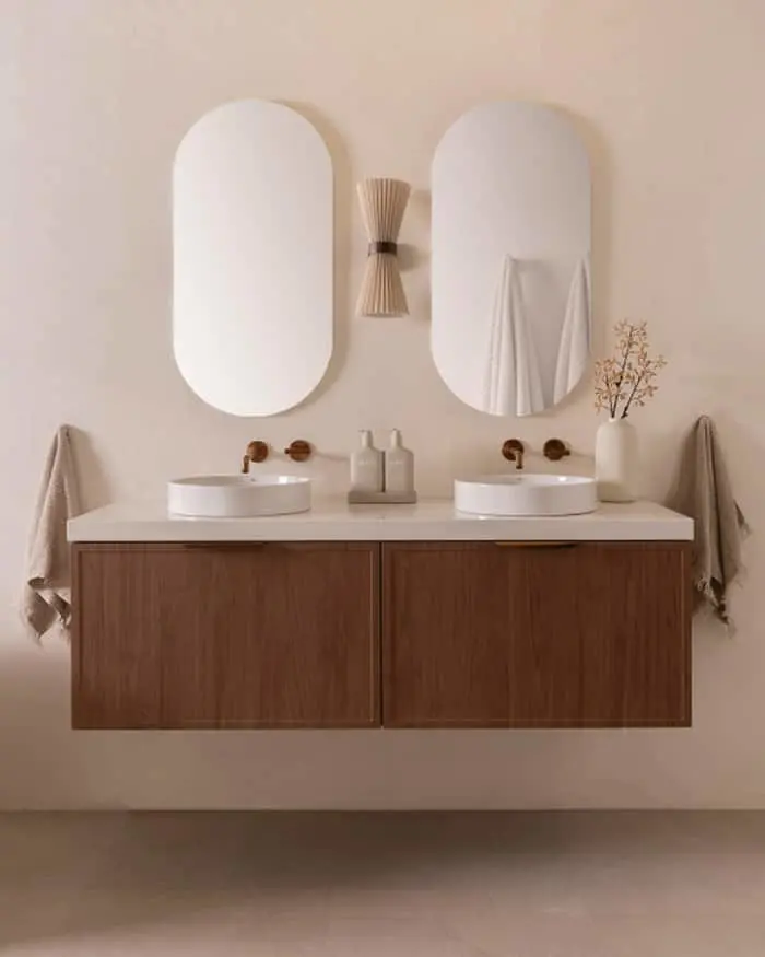 Modern bathroom vanity with wooden cabinets, white countertop, dual sinks, oval mirrors, and minimal decor in neutral tones.