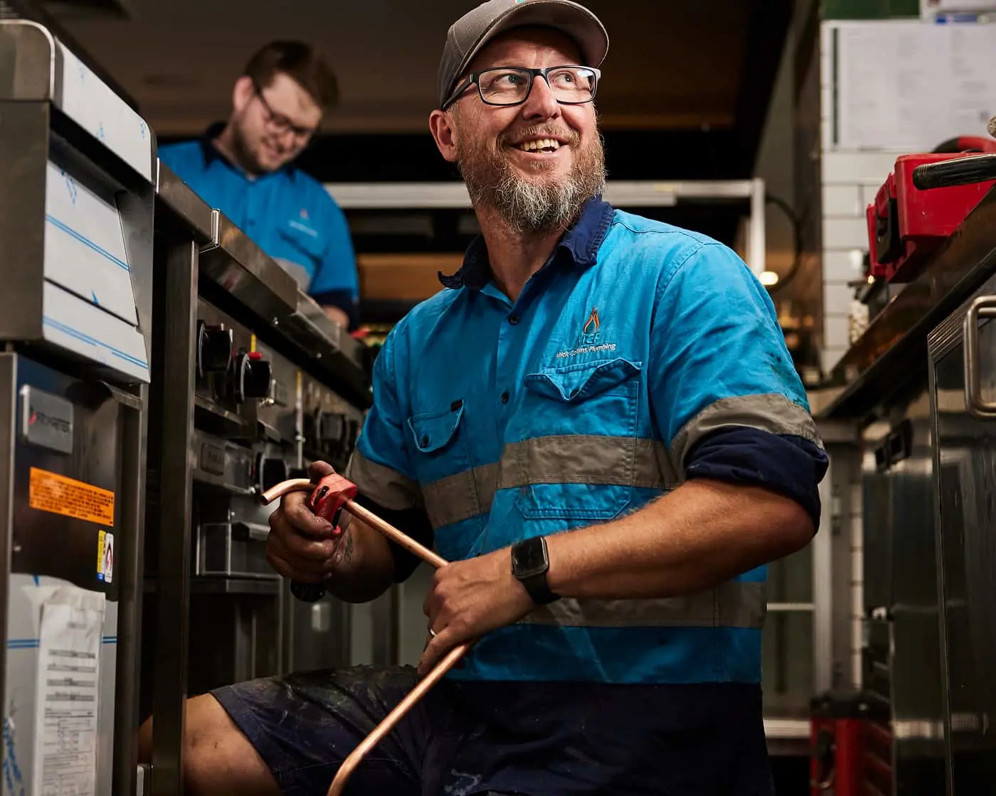 Smiling tradesperson in blue workwear and cap holding copper pipe in workshop with colleague in background.
