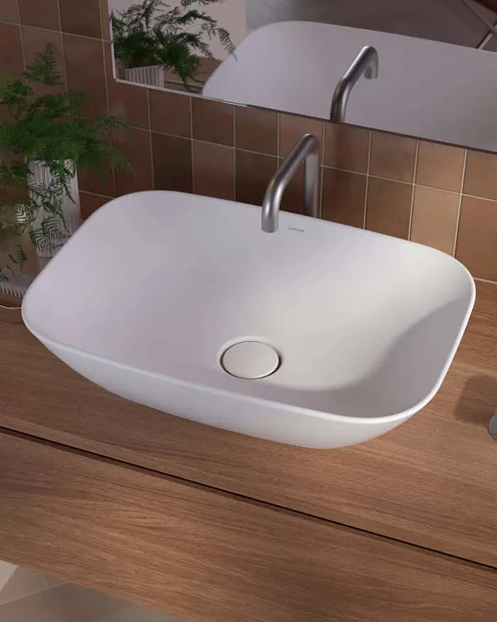 Modern white vessel sink with stainless steel faucet on wooden vanity, brown tiled wall and green plant visible.