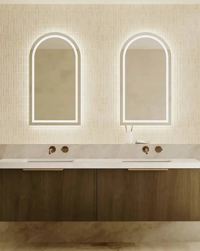 Modern bathroom with two arched backlit mirrors above a floating dark wood vanity with white sinks and brass fixtures.