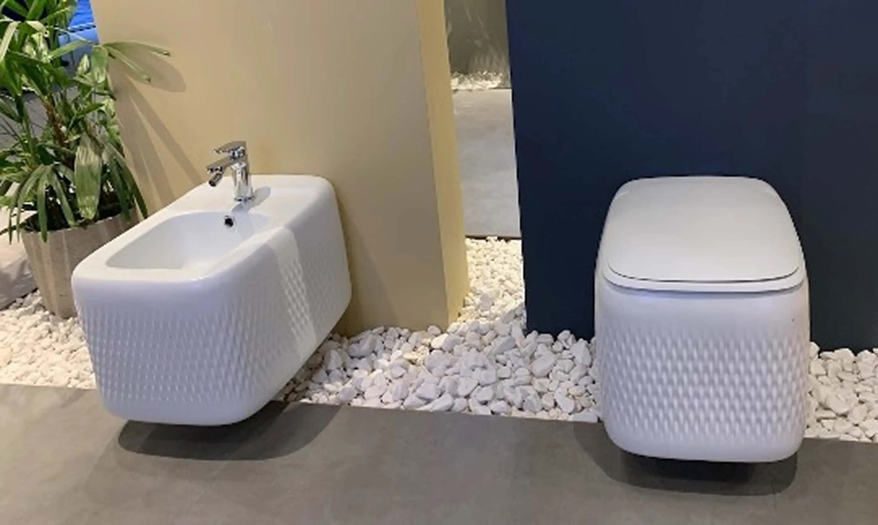 Modern white bidet and toilet with textured design on a bed of white pebbles, with a green plant in the corner.