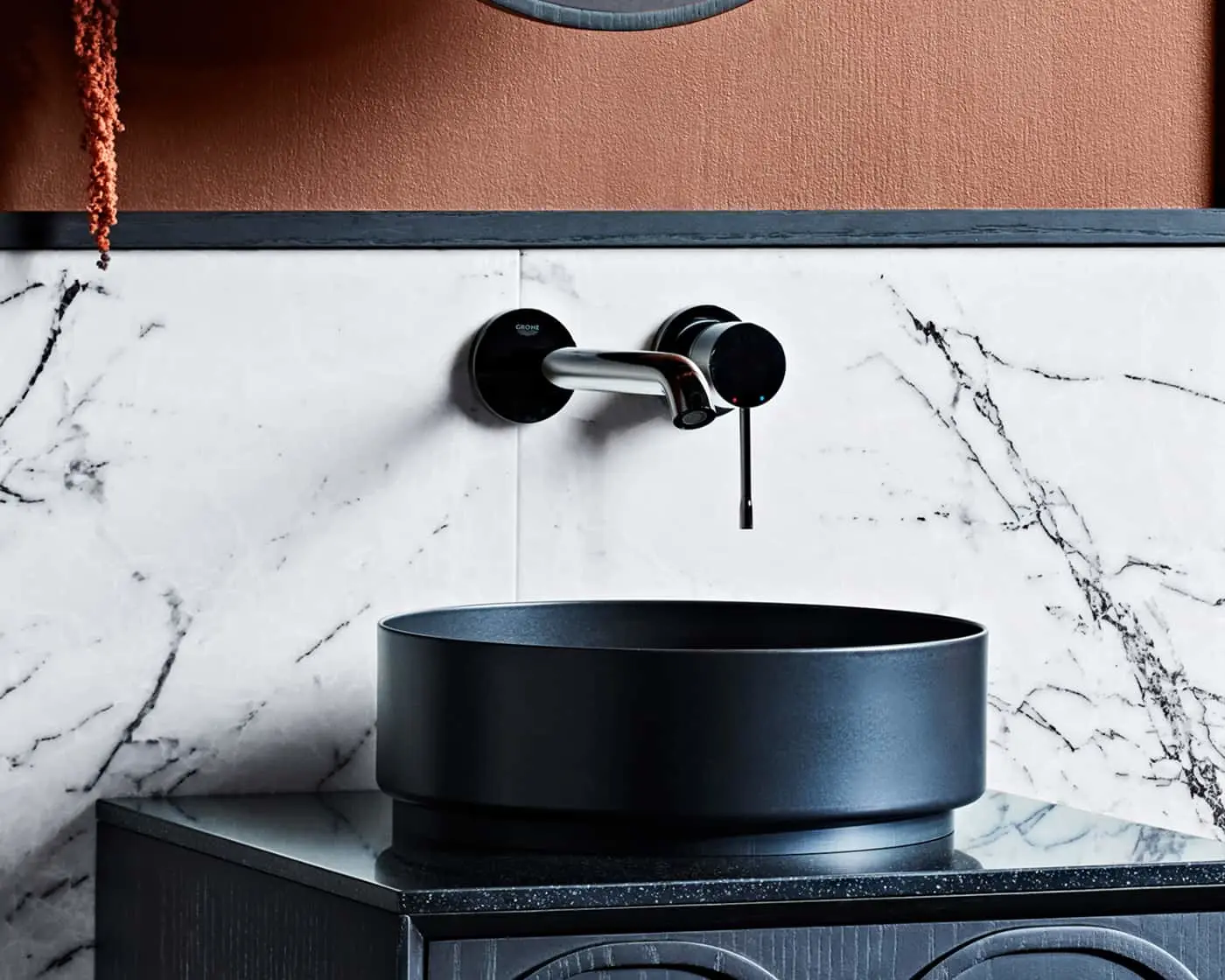 Modern black vessel sink with wall-mounted chrome faucet against white marble backsplash and terracotta wall.