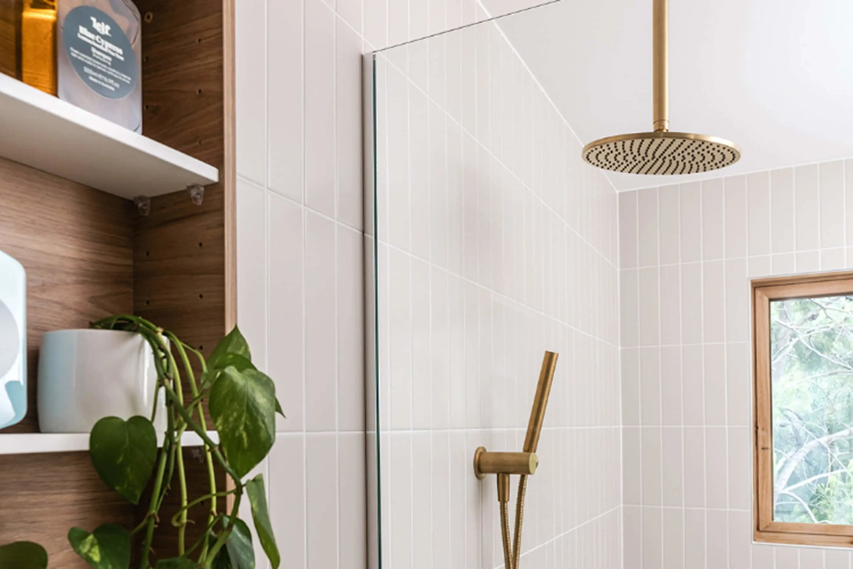 Modern bathroom with white tile walls, brass rainfall showerhead, glass partition, wooden shelving with plants, and window.