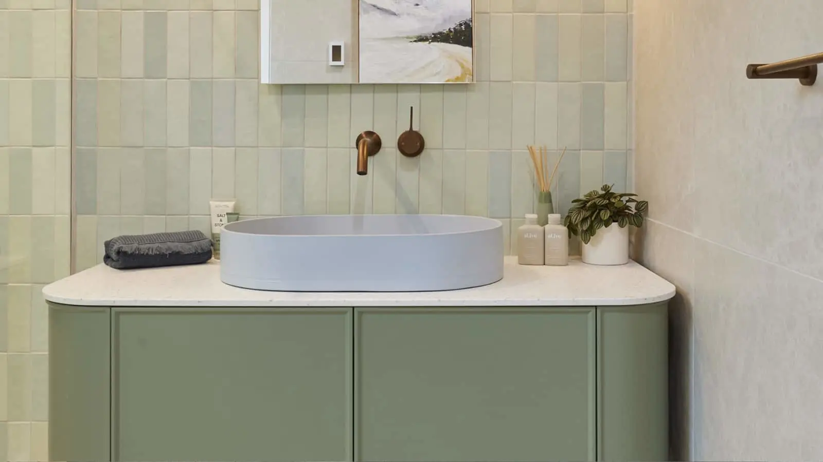Modern bathroom with light blue vessel sink on sage green vanity, brass fixtures, and mint tile backsplash.