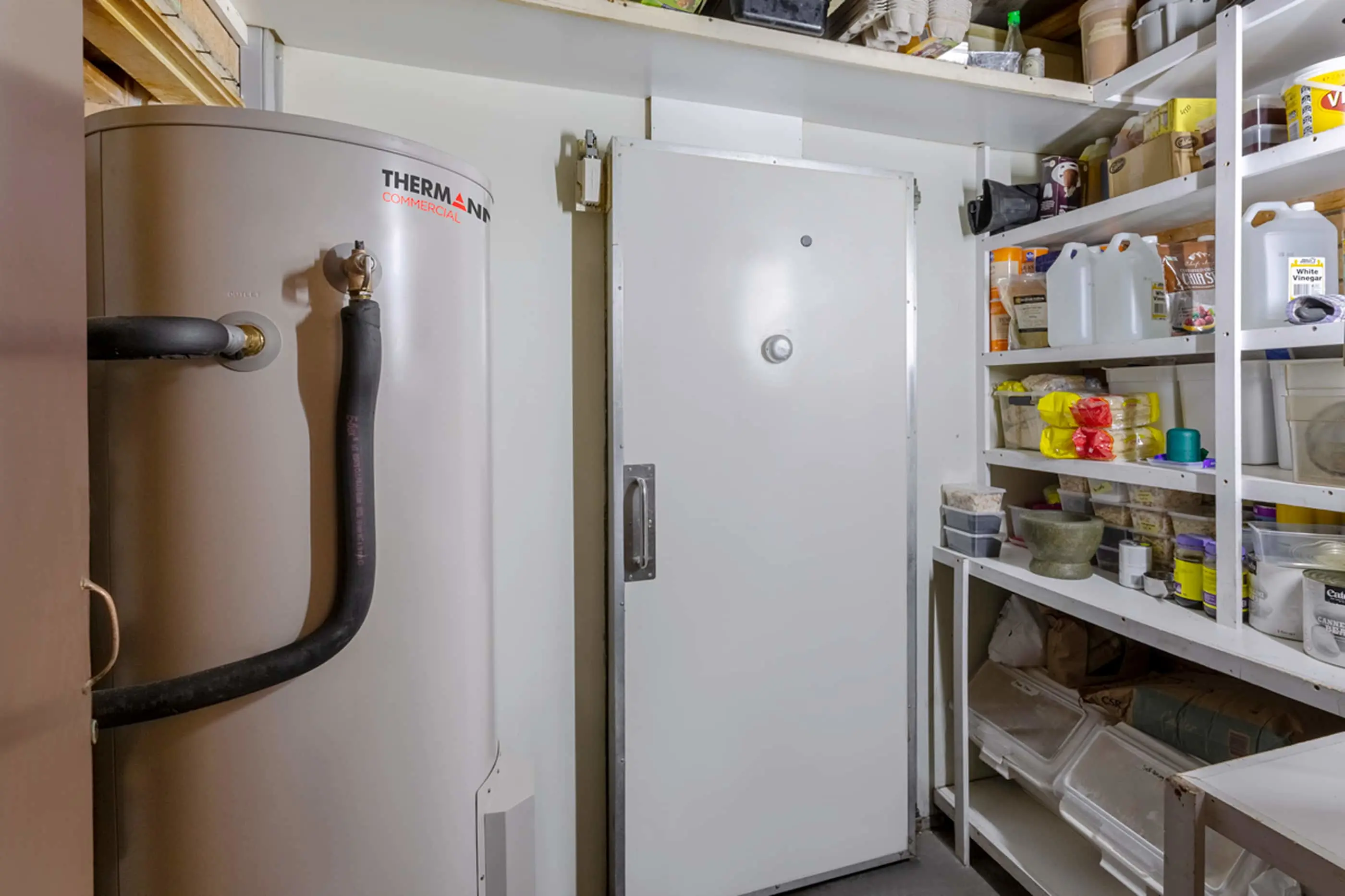 Utility room with Thermann water heater, white cold storage door, and shelving stocked with household supplies and containers.