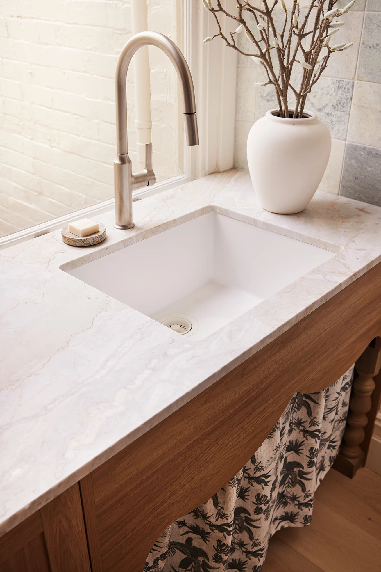 White undermount kitchen sink with brushed nickel faucet set in a marble countertop on a wood cabinet, with a white vase nearby.