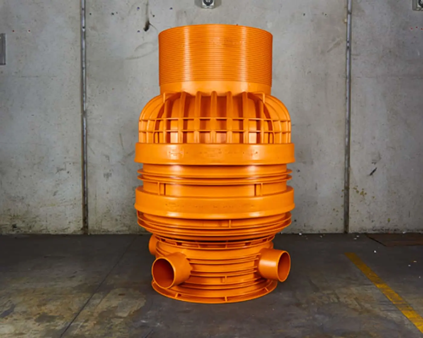 Bright orange industrial pipe connector with ribbed sections against a concrete wall background.