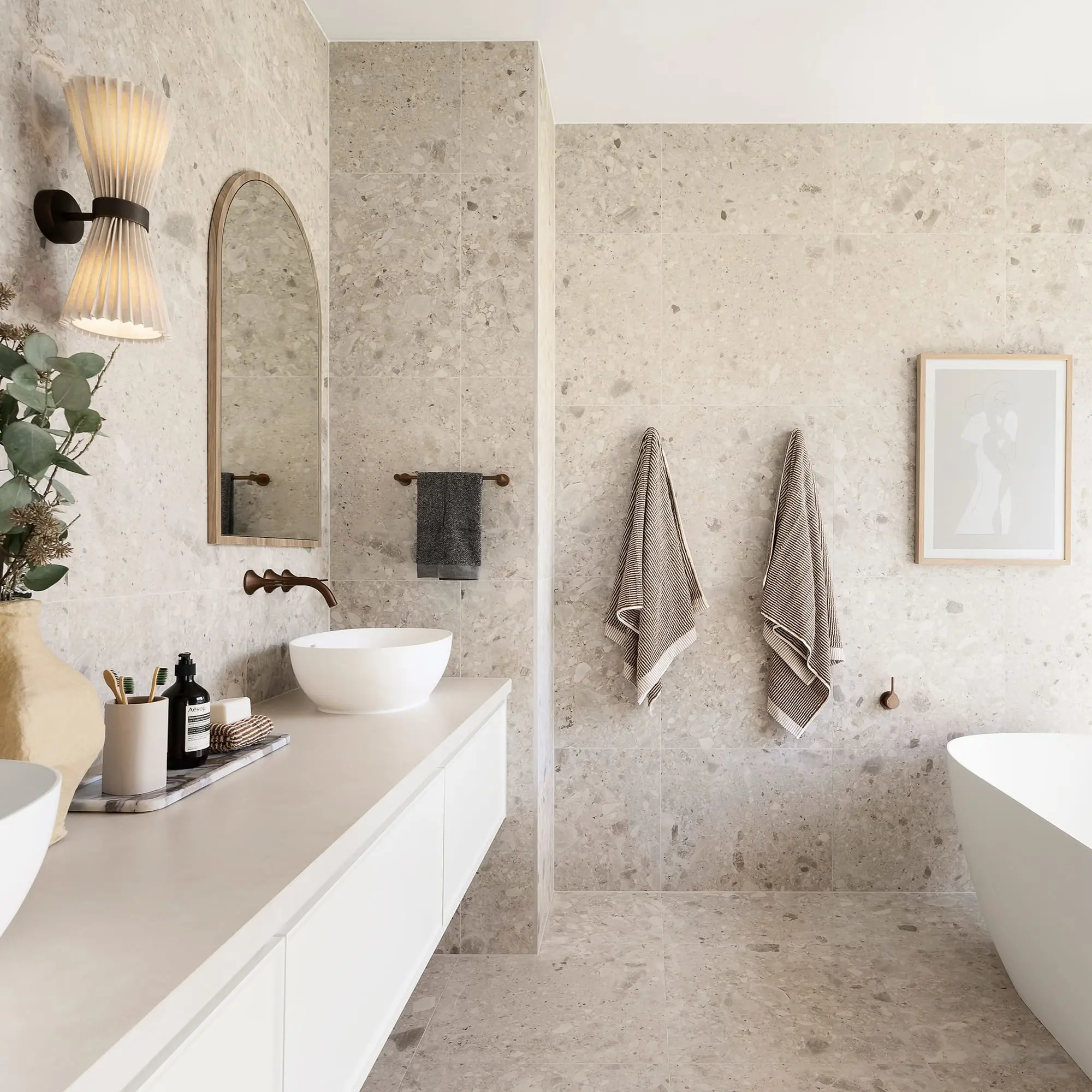 Modern bathroom with stone walls, white vanity, vessel sink, arched mirror, wall sconce, hanging towels, and partial bathtub view.