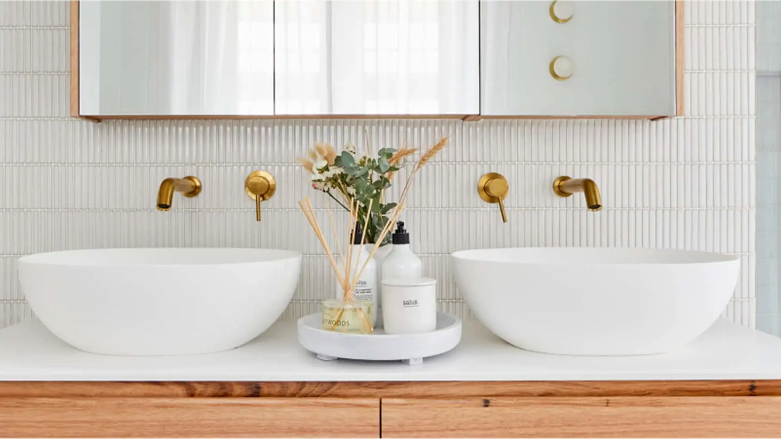 Modern bathroom with dual white vessel sinks, brass wall-mounted faucets, and decorative items on a round tray.