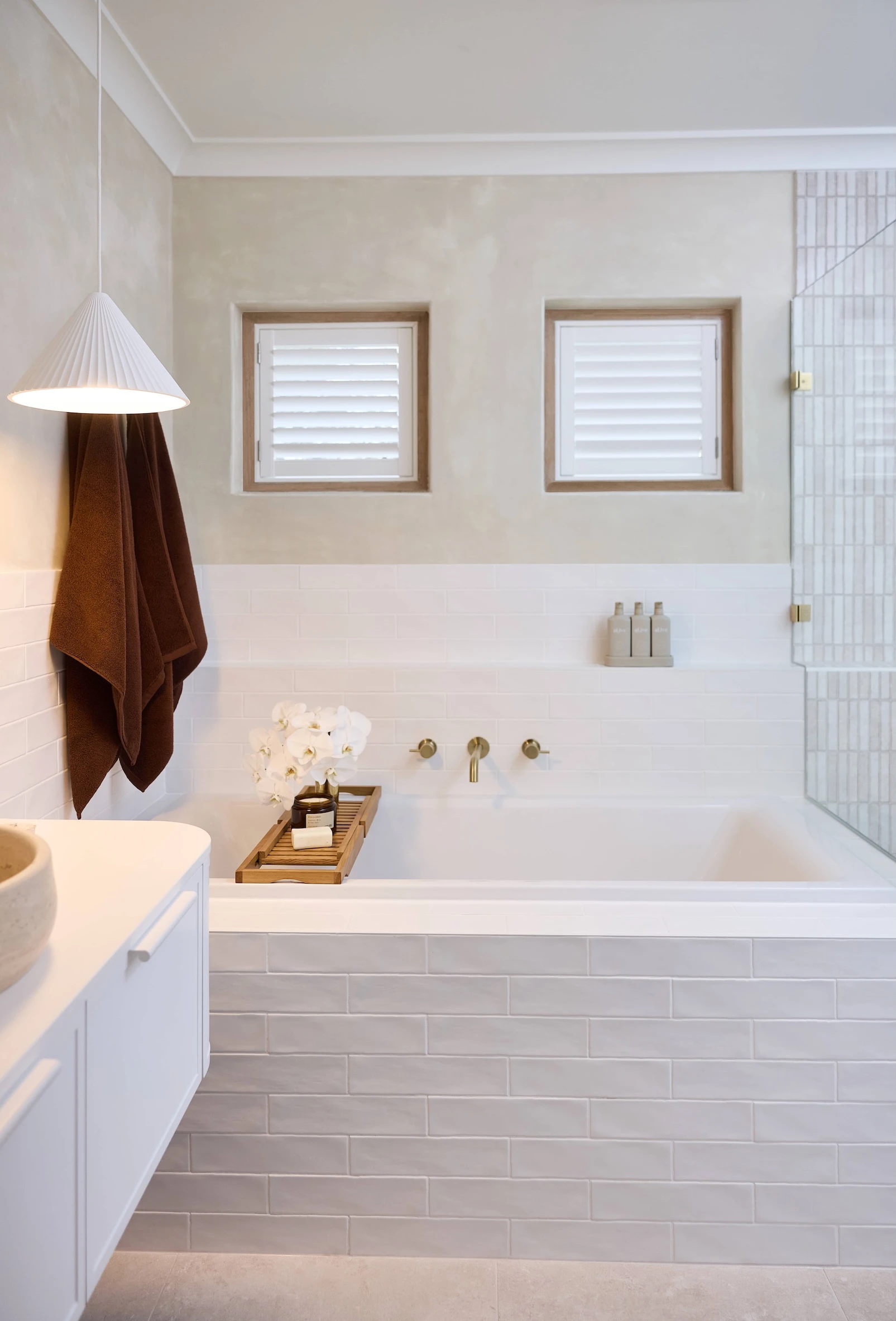 Modern bathroom with white bathtub, brown towel, pendant light, two small windows with shutters, and tiled walls.
