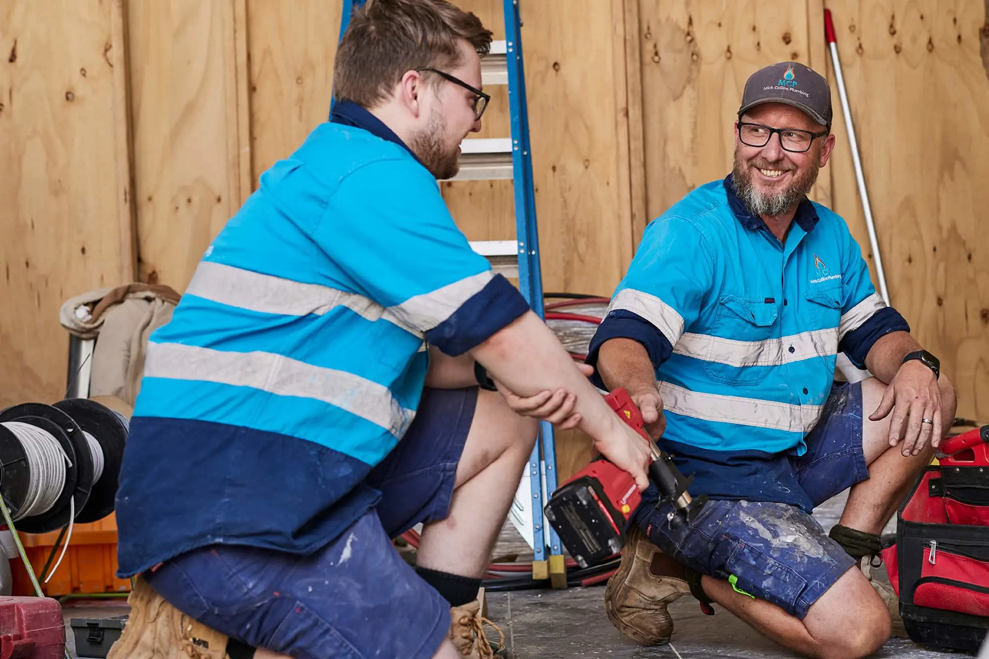 Two workers in blue hi-vis shirts with reflective stripes conversing at a construction site with tools and a ladder nearby.