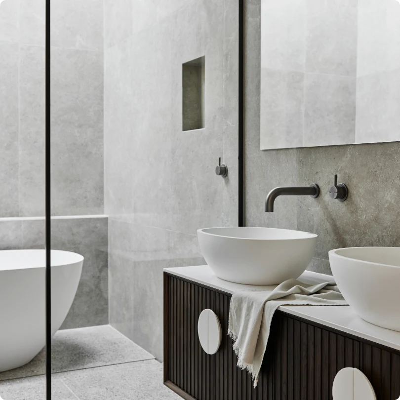 Modern bathroom with gray walls, white vessel sinks on dark wood vanity, and freestanding tub behind glass partition.