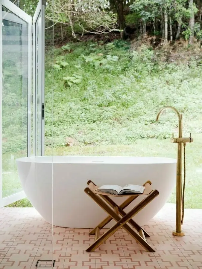 White freestanding bathtub with brass fixtures and wooden stool holding open book, overlooking forest through glass walls.