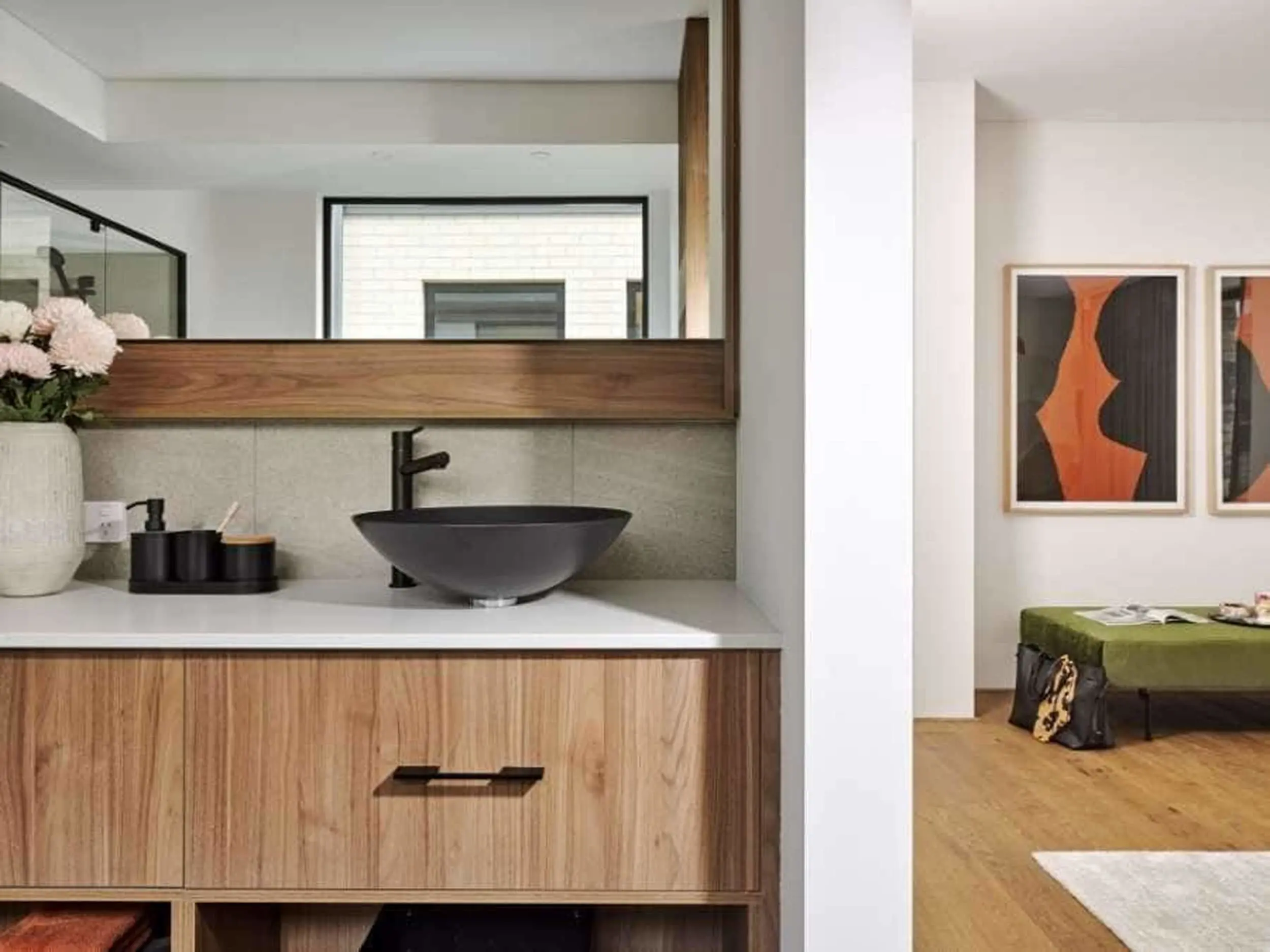 Modern bathroom with black vessel sink, wooden vanity, large mirror, and abstract artwork visible in adjacent room.