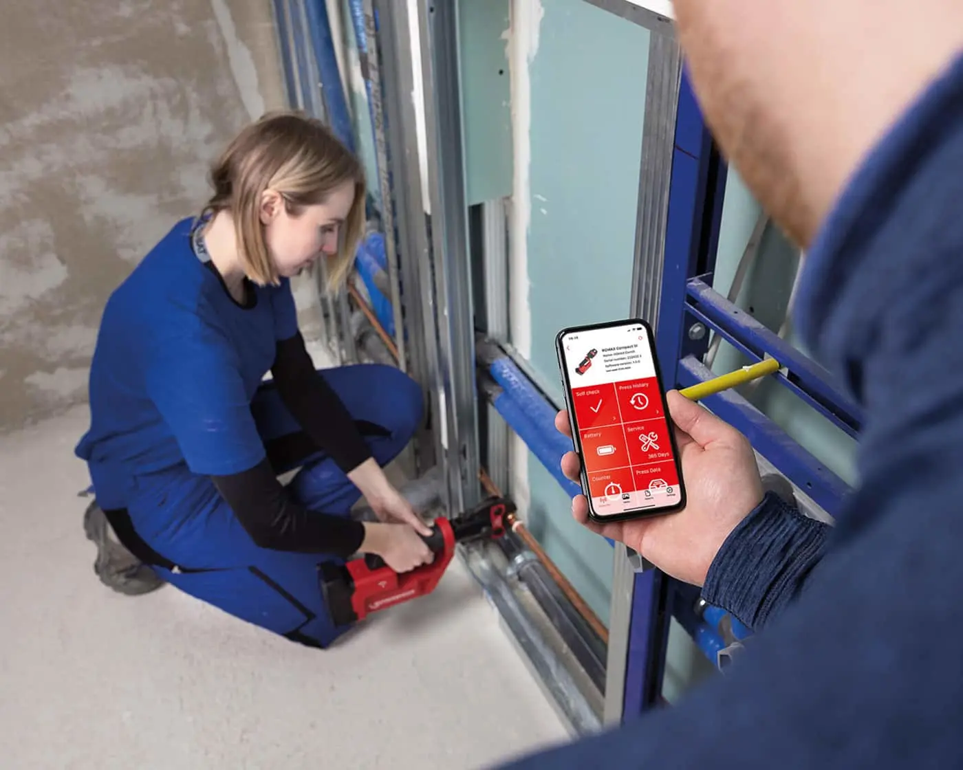 Person in blue workwear using red tool on metal framing while another checks mobile app with red interface